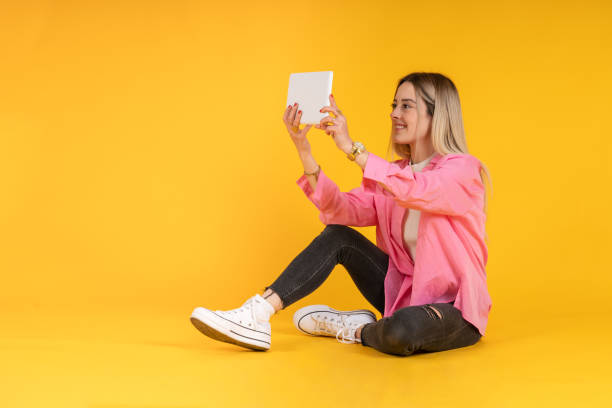 seated young woman making a video call with a tablet dressed in black jeans and a pink shirt - shirt women pink jeans imagens e fotografias de stock