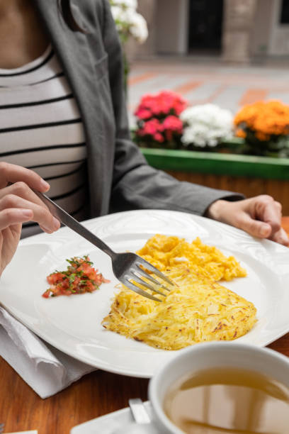 restaurant table with served plate with a fresh breakfast, scrambled eggs and cup with tea, details of hands holding a fork