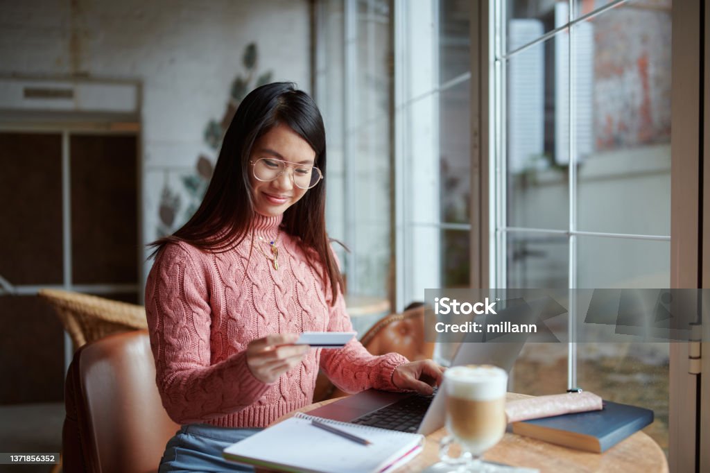 A happy Asian girl sitting in the cafeteria and using her credit card for online shopping on the laptop. Online Shopping Stock Photo