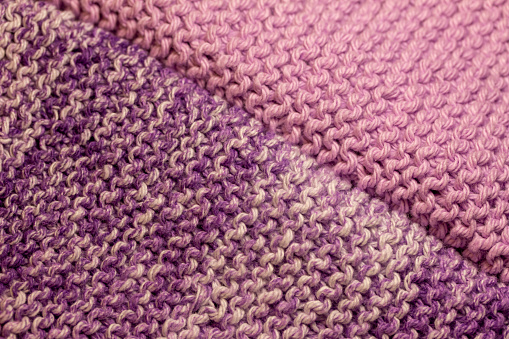 This image shows a macro abstract texture background of two hand-knitted yarn swatches in a basic garter stitch, in varying shades of lavender purple and white.