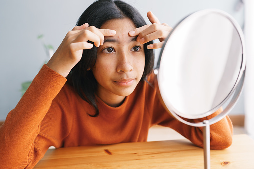 Joven asiática Exprimiendo la piel del acné y mirándose en el espejo en casa photo