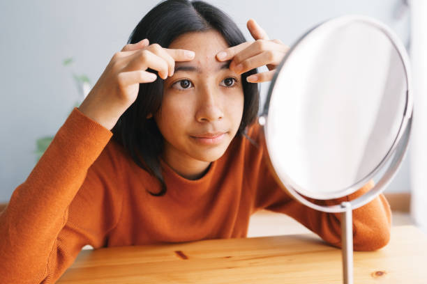 Asian young woman Squeezing acne skin and looking on the mirror at home stock photo