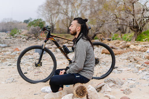 young latino male long haired hispanic athlete sitting in a park in the morning with his bicycle on the side while he is relaxing meditating. side view