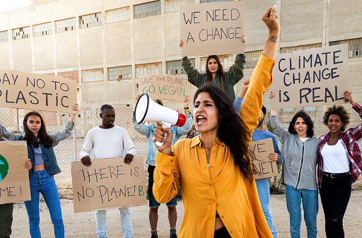 Young Caucasian woman with arm raised shouting through megaphone leads demonstration protesting against global warming and plastic pollution. Protesters holding placards in background. Activism.
