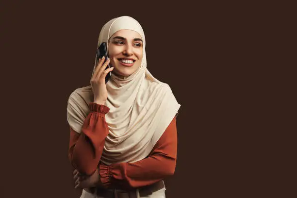 Beautiful muslim woman dressed in beige hijab holding phone smiling and talking on brown background. Copy space.