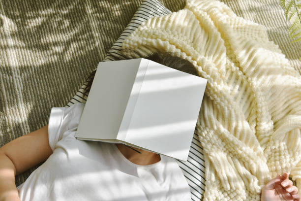 女性は本を読みながら眠りに落ち、日当たりの良い怠惰な日に柔らかく快適なニット毛布で休んで冷やしている女の子は、アジアの女性が床に横たわっている本で彼女を覆います。 - oversleep ストックフォトと画像