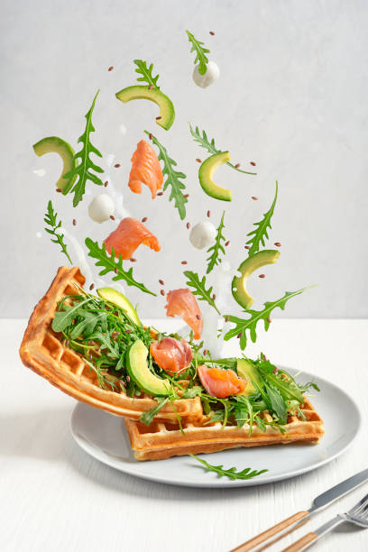Belgian waffles with levitating ingredients of arugula, salmon, avocado and mozzarella cheese served on plate on table Belgian waffles eaten as a breakfast food with topping of fresh green arugula leaves, avocado slices, mozzarella cheese and salmon levitating or falling on plate served on white wooden table arugula falling stock pictures, royalty-free photos & images