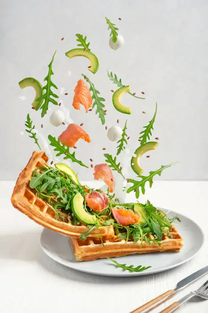 Photo of Belgian waffles with levitating ingredients of arugula, salmon, avocado and mozzarella cheese served on plate on table