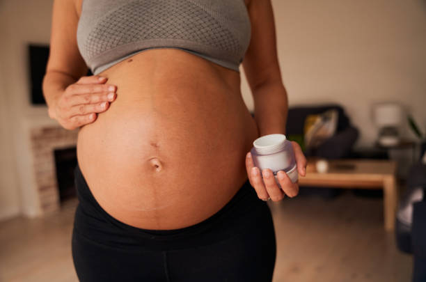 torso de la mujer caucásica embarazada de pie, aplicando crema en su vientre. estilo de vida feliz y activo en el hogar moderno durante el día - ewan fotografías e imágenes de stock