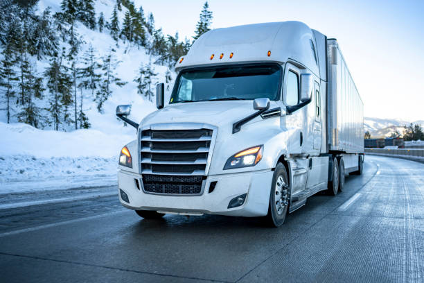 masknet big rig biała naczepa z włączonymi reflektorami przewożąca ładunek w suchej naczepie furgonetkowej poruszającej się po zimowej górskiej drodze z dużą ilością śniegu i lodu na śliskiej nawierzchni - semi truck vehicle trailer truck empty zdjęcia i obrazy z banku zdjęć