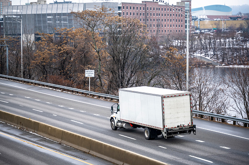 Middle power industrial day cab semi truck with long box trailer transporting commercial cargo running on the divided winter highway road along the river in Manchester city