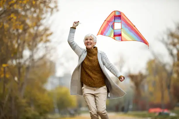 Happy mature woman having fun while running with a kite during autumn day in the park. Copy space.