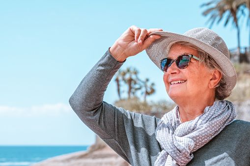 Smiling youthful granny in outdoors at sea enjoying sunny day and vacation, holding her hat looking at horizon over water. Senior carefree woman enjoying retirement and freedom