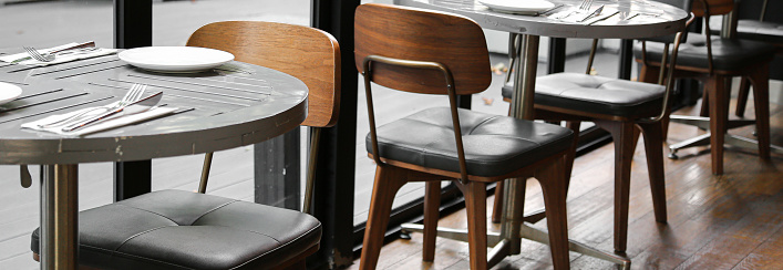 Tableware on round gray plank table with metal base and wood chair with black cushion on old wooden flooring near black frame ceiling to floor window empty retro cafe interior design