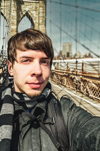Young adult man with brown hair and beard taking a selfie, looking at camera, standing on brooklyn bridge new york nyc, usa