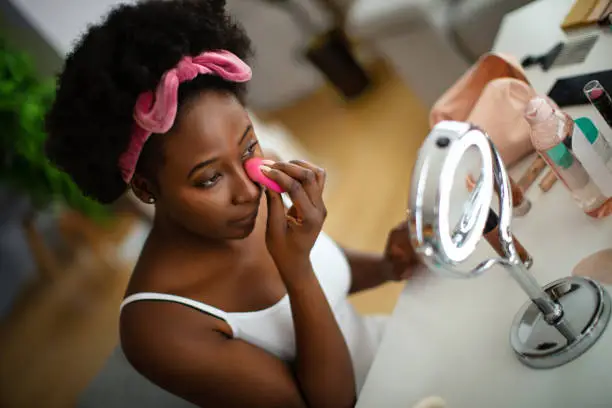 Beautiful young woman applying concealer on her face with a beauty blender, starting with her beauty routine.