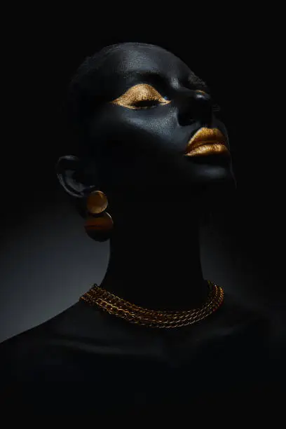 Beauty woman painted in black skin color body art, gold chain in his hands and around his neck. Gold makeup lips eyelids, fingertips nails in gold color paint. Professional makeup