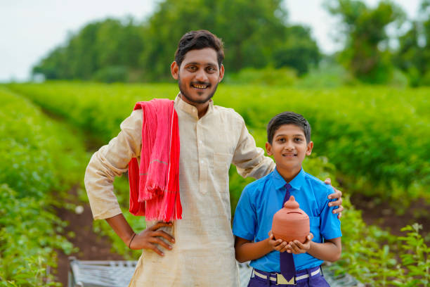 importance of saving concept : Smart indian little boy standing and holding piggy bank in hand with his father at agriculture field. stock photo
