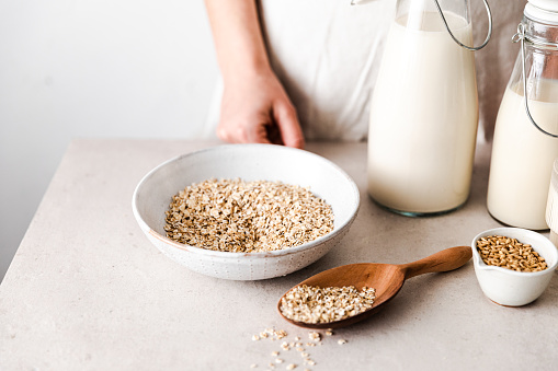 Close-up of a woman preparing oat milk at home. Bowl of oat flakes with a wooden scoop and two bottles full of oat milk.