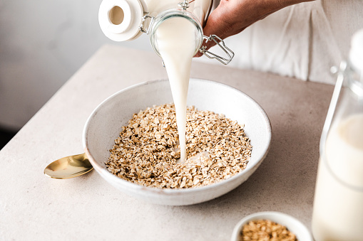 Close-up of pouring milk into oat flakes bowl on table. Preparing healthy vegan breakfast.