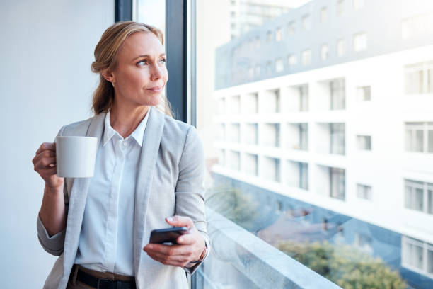 aufnahme einer reifen geschäftsfrau, die aus einem fenster schaut, während sie ein smartphone benutzt und in einem modernen büro kaffee trinkt - looking through window window business women stock-fotos und bilder