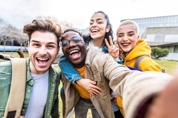 Multiracial students company taking selfie portrait in university campus - Multi ethnic best friends laughing at camera outside - Teens having fun together - Youth culture and school concept stock photo