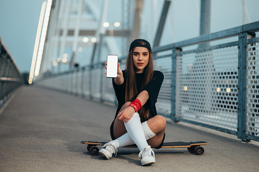 Young woman sitting on her longboard and showing a smartphone while on the bridge