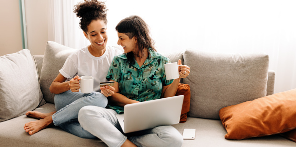 Enjoying the online weekend discounts. Happy young lesbian couple shopping online while relaxing together in their living room at home. Smiling female lovers spending quality time together.