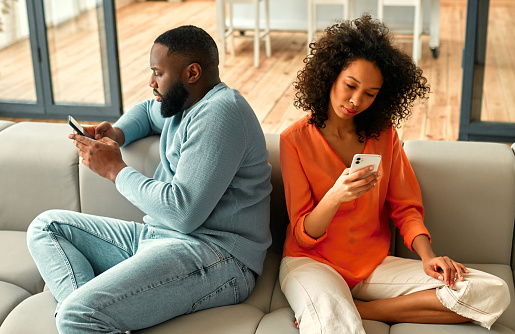 African american couple sitting on sofa with phones facing away from each other in living room at home.