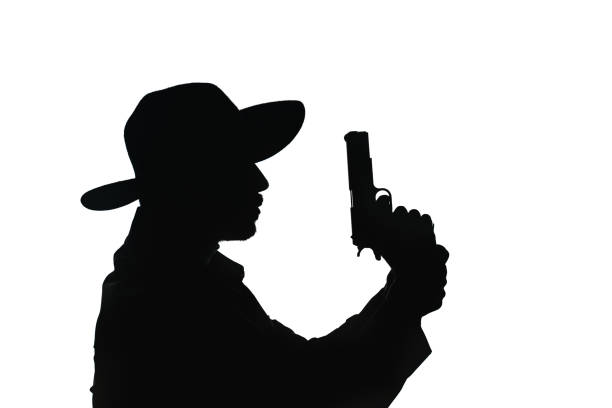 Man wearing fedora hat aiming gun with a furious face.The focus is on the model's face.Gun is slightly blurred.The photo is black and white.Shot in horizontal framing with a full frame DSLR camera.Background is black.He is wearing a waist and shirt.