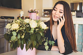 美しいアジアの女性は、白いバラの花の花束を賞賛し、明るいインテリアのリビングルームのテーブルのそばに座って電話を話します。笑顔の若い女性は、驚きの贈り物を受け取って幸せ。