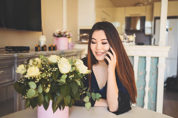 Beautiful Asian woman speaks phone sits by the table in living room bright interior admires white roses flowers bouquet. Smiling young lady happy to receive a surprise gift. stock photo