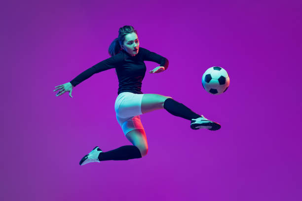 ein sportliches mädchen, eine fußballspielerin kickt im sprung fußballball isoliert auf lila studiohintergrund in neonlicht. sport, action, bewegung, fitness - exercising women sport studio shot stock-fotos und bilder