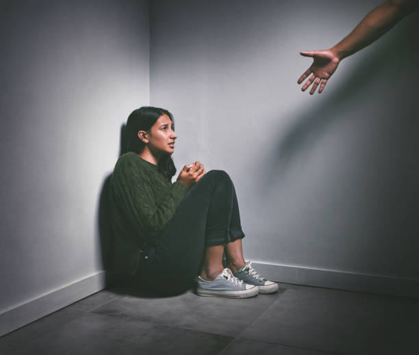 photo d’une jeune femme assise dans le coin d’une pièce sombre avec une main tendue pour l’aider - désespoir photos et images de collection