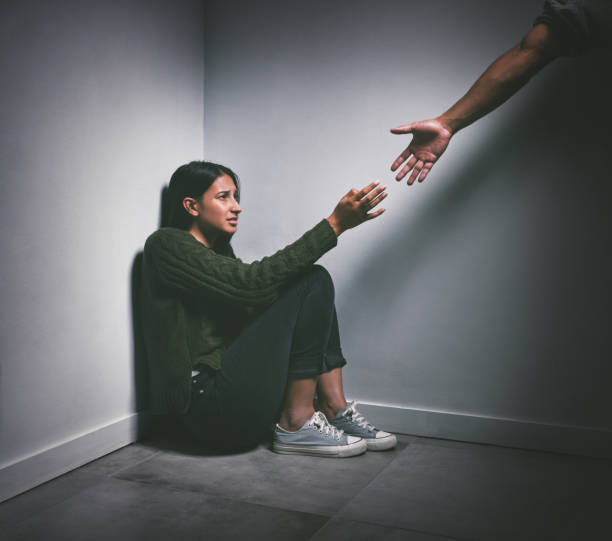 foto de una joven sentada en la esquina de una habitación oscura con una mano extendida para ayudarla - drug abuse addiction women violence fotografías e imágenes de stock