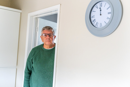 Senior Caucasian man peering around doorway at home with clock on wall showing two minutes to twelve