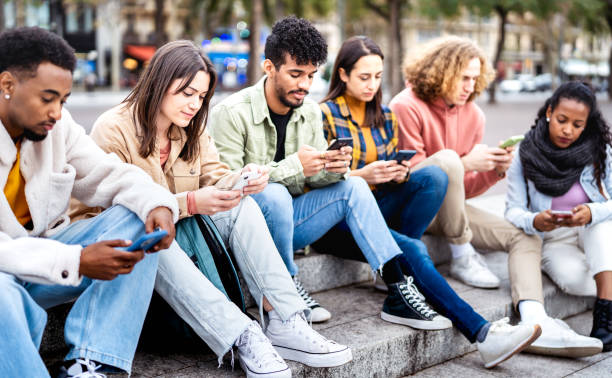 Bored friends group using mobile smart phone sitting at university college yard - Young people addicted by smartphone devices - Technology concept with always connected students on bright vivid filter stock photo