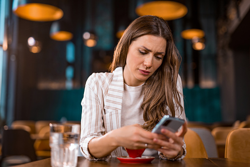 Worried and displeased young woman reading bad news on her phone while sitting at coffee shop.