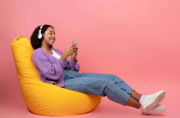 mujer negra con auriculares y teléfono inteligente escuchando música, disfrutando de audiolibros, sentada en una silla de bolsa de frijoles - chill out audio fotografías e imágenes de stock