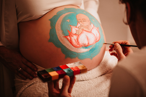 Close up of the father-to-be painting the pregnant belly of his beloved wife painting a baby girl sitting in a lotus in the centre of a mandala. Color editing and added grain. Real life situation. Part of a series.