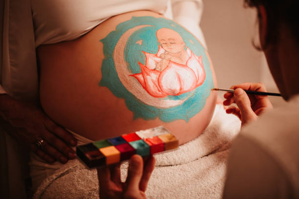 Close up of the father-to-be painting the pregnant belly of his beloved wife painting a baby girl sitting in a lotus in the centre of a mandala stock photo