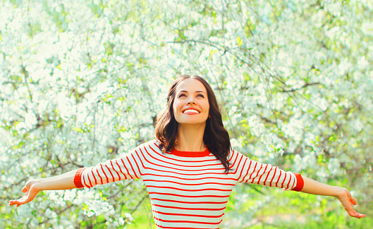 Happy smiling young woman raising her hands up in spring blooming garden on flowers background