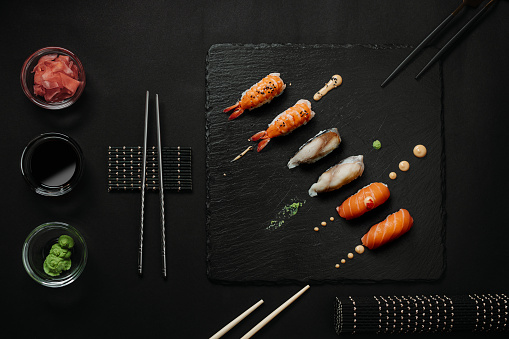 Table top view of salmon, shrimp and mackerel nigiri and decorative sauce drops on a black stone plate on black background, with chopsticks and three glass bowls containing soy sauce and green wasabi