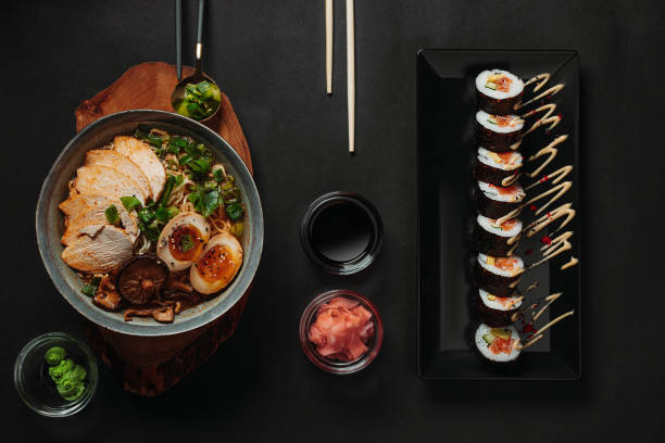 갈색 나무 접시에 파란 그릇에 라멘의 테이블 상단 보기와 간장, 절인 생강, 와사비, 황금 숟가락에 파와 어두운 배경에 젓가락검은 유리 접시에 초밥 - hosomaki 뉴스 사진 이미지