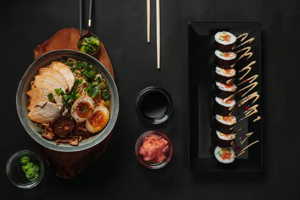 Photo of Table top view of ramen in a blue bowl on a brown wooden plate and sushi on a black glass plate with soy sauce, pickled ginger, wasabi, scallions in a golden spoon and chopsticks on a dark background