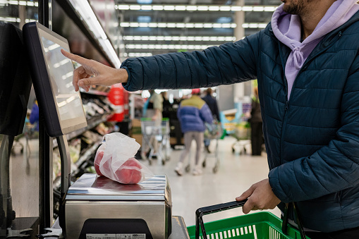Side view of a man shopping in a supermarket while on a budget. He is weighing the items at the self service checkout in the North East of England. The peppers are in a sustainable mesh bag.