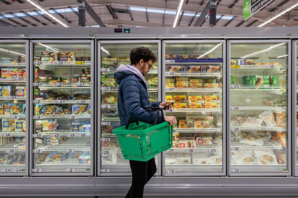 Buying Convenient Food Man shopping in a supermarket while on a budget. He is looking for low prices due to inflation, standing looking at his phone in front of a row of freezers. He is living in the North East of England. retail stock pictures, royalty-free photos & images