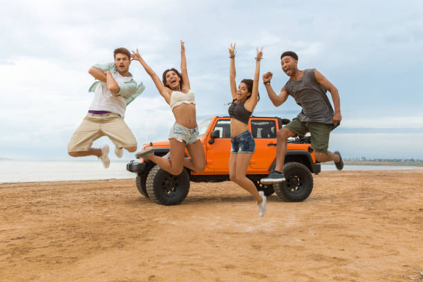Group of happy multiethnic friends jumping in front of their car with hands raised and having fun at the beach