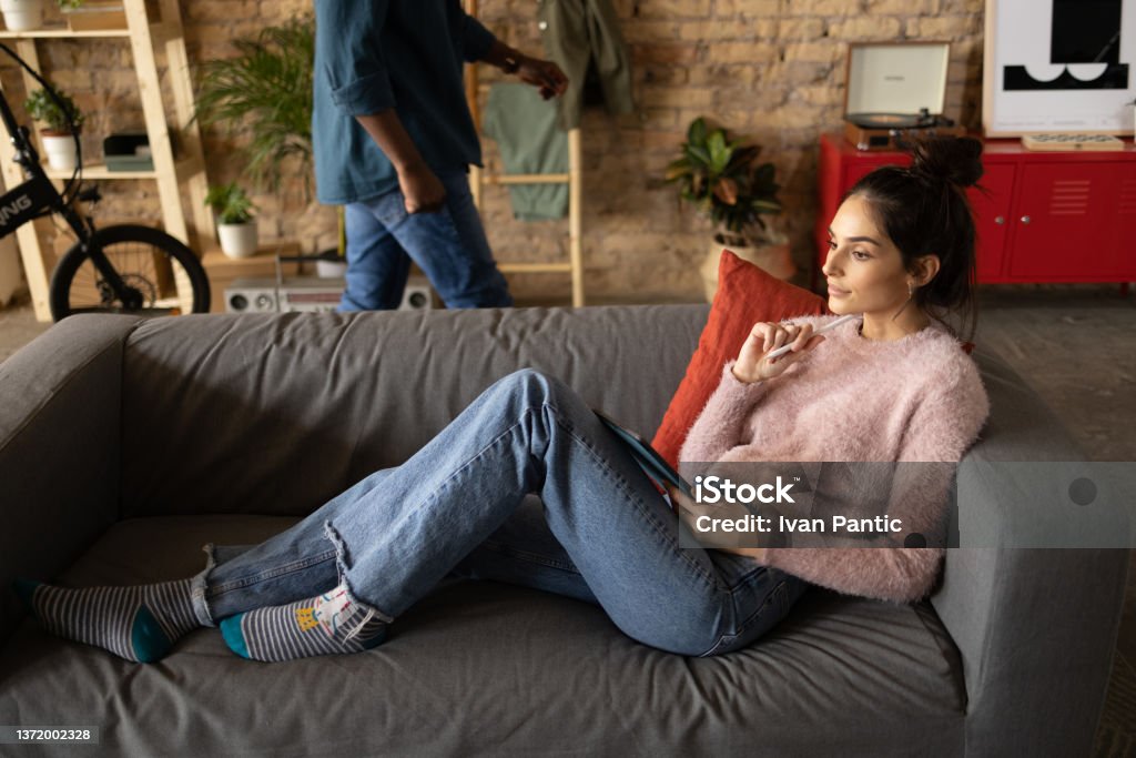 Young Caucasian woman relaxing at home Young attractive Caucasian woman relaxing in her loft apartment, resting on a sofa in the living room and holding a digital tablet computer in her hand Roommate Stock Photo