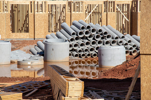A stack of concrete pipes is reflected in stormwater accumulated in an excavated area of a construction site.  Stacked wooden trusses in foreground, chipboard clad building in background.